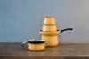 A set of orange pots and pans stacked on a wooden table against a textured blue-gray wall. Podgląd zdjęcia produktu