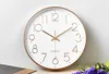 A white wall clock with a gold frame and gold numbers is displayed against a light gray and white background. Produktbild-Vorschau