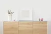 A white framed picture with a light grey insert sits on top of a light wood cabinet. To the left of the frame is a white vase with dried flowers and a small pink and white bowl. To the right of the frame are two small decorative objects, one green and one  Náhled obrázku produktu