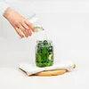 A hand is pouring liquid from a glass into a jar filled with pickles. Produktbild-Vorschau