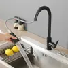 A hand is holding a black pull-down kitchen faucet spray head, with water spraying into a stainless steel sink. Two lemons are visible in a drying rack in the sink. Two spice grinders are in the background. Produktbild-Vorschau