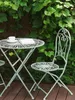 A metal bistro set with a round table and two chairs is set up on a wooden deck, surrounded by lush green foliage. Náhľad obrázku produktu