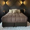 A bed with brown bedding and decorative pillows, set against a dark, geometric wall with wall sconces. Produktbild-Vorschau
