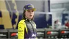 A woman in racing attire stands in front of a blurred background. Produktbild-Vorschau