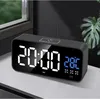 A digital alarm clock is displayed on a surface, with a window and a plant in the background. Podgląd zdjęcia produktu