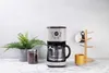 A coffee maker with a carafe, a mug tree with mugs, a yellow mug, a jar of coffee beans, and a potted plant on a marble countertop. Náhled obrázku produktu