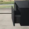 A close-up view of a black bathroom vanity with one drawer partially open, showing the interior. The background features a blurred outdoor scene with greenery and a concrete floor. Produktbild-Vorschau
