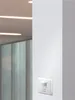 A white motion sensor is mounted on a white wall next to a blurred background of a modern office interior with ceiling lights. Produktbild-Vorschau