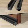 Two black door draft stoppers are shown on a wooden floor, with one partially installed on a wooden door frame. Náhled obrázku produktu