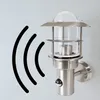 A modern outdoor wall lamp with a motion sensor is mounted on a light gray wall. Black wavy lines indicating motion detection emanate from the left side of the lamp. Produktbild-Vorschau