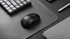 A black wireless mouse is placed on a dark gray mousepad, next to a keyboard and a stylus pen. Produktbild-Vorschau