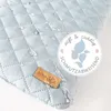 A close-up of a light blue quilted surface with water droplets, featuring a leather tag and a circular marketing graphic. Anteprima dell’immagine prodotto