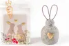 Two Easter-themed decorative items: a white paper bag with a carrot clip and bunny graphics, and a gray ceramic bunny bell with a gold heart. Produktbild-Vorschau