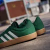 A pair of green Adidas sneakers with white stripes and a gum sole, resting on a concrete surface with a blurred urban background. Produktbild-Vorschau