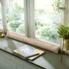 A beige fabric draft stopper is placed on a windowsill in front of a window with a garden view. Books are stacked on the windowsill in front of the draft stopper. Produktbild-Vorschau