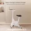 A white stationary exercise bike with a laptop on its adjustable tray, set against a neutral background with decorative elements on the left. Voir les images du produit
