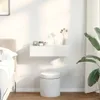 A white floating shelf with two drawers is mounted on a white wall above a white cylindrical stool. Decorative items and a vase with dried flowers are on the shelf. A bed and a large plant are partially visible. Náhľad obrázku produktu