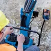 A person wearing a yellow hard hat and orange safety vest is operating a blue mini-excavator. Produktbild-Vorschau