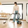A woman is vacuuming a wooden floor in a bright, modern living room with large windows. Náhled obrázku produktu