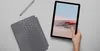 A Microsoft Surface Go 2 tablet, keyboard cover, and stylus are laid out on a light gray surface. A hand is holding the tablet. Produktbild-Vorschau