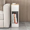 A narrow white nightstand with a wooden top is placed next to a white sofa. The nightstand has a drawer and an open shelf displaying several books. Produktbild-Vorschau
