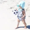 A baby on a sandy beach holding a toy soccer ball. Produktbild-Vorschau