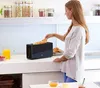 A woman is putting a piece of bread into a black toaster on a kitchen counter. Produktbild-Vorschau