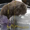 A gold chain necklace is displayed on a reflective surface with various crystals and rocks in the background. Produktbild-Vorschau