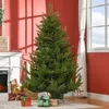 A tall, green artificial Christmas tree stands in a room with red walls, near a fireplace and a window with white curtains. Náhled obrázku produktu