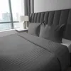 A neatly made bed with gray striped satin bedding and two pillows, set against a dark gray tufted headboard, with a large window and cityscape visible in the background. Produktbild-Vorschau