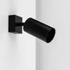 A black cylinder-shaped spotlight is mounted on a white wall. Produktbild-Vorschau