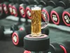 A beer mug with a dumbbell-shaped handle sits on top of a dumbbell, with a rack of dumbbells in the background. Produktbild-Vorschau