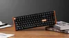 A black keyboard with orange accents is placed on a wooden desk next to a notebook and some books. Voir les images du produit