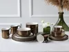 A collection of bronze and white ceramic dishes, including mugs, bowls, and plates, arranged on a white surface. Náhľad obrázku produktu