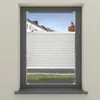 A window with white pleated blinds partially covering the view of houses and a tree. Produktbild-Vorschau