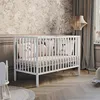 A white baby crib with pillows inside, set in a room with patterned wallpaper and curtains. Anteprima dell’immagine prodotto