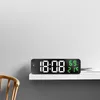 A digital clock is placed on a white shelf against a light gray wall. A wooden chair is partially visible on the left. Voir les images du produit