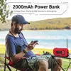 A man is sitting in a camping chair outdoors, looking at his phone, with a red radio and power bank on a table next to him. The background shows a lake and trees. Produktbild-Vorschau