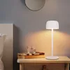 A white LED table lamp sits on a wooden nightstand next to a bed, with books and a glass of water. Produktbild-Vorschau