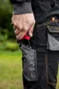 A person's hand is reaching into the pocket of black work pants, holding a red and black tool. Produktbild-Vorschau