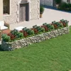 A gabion raised garden bed filled with plants is situated on a wooden deck next to a stone building and a grassy lawn. Náhled obrázku produktu