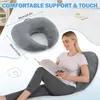 A woman is sitting on a bed, reading a book, and leaning against a large U-shaped pillow that is also shown separately with dimensions. The pillow is designed for support while gaming or reading. Produktbild-Vorschau