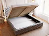 A bed with its storage compartment open, showing the slatted base and interior storage space. The bed is upholstered in gray fabric with a tufted headboard and frame. The scene is set in a bedroom with wooden flooring and a window in the background. Produktbild-Vorschau
