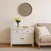 A clock on a wall above a white cabinet with flowers and a book, next to a sofa. Produktbild-Vorschau