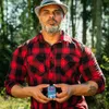 A man in a lumberjack shirt and hat holds a small tin of product in his hands, outdoors in a wooded area. Náhľad obrázku produktu