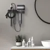 A bathroom shelf with a hair dryer holder mounted on the wall, holding a hair dryer and its accessories. A plant and various bottles are on the shelf below. Produktbild-Vorschau