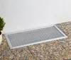 A rectangular metal grate cover with a grid pattern is placed on a gravel path next to a white textured wall. Produktbild-Vorschau