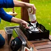 A person is pouring liquid from a bottle into a metal burner on a wooden table outdoors. Produktbild-Vorschau