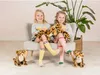 Two young girls sitting on a chair, holding stuffed leopards, with stuffed leopards on the floor. A large plant is in the background. Produktbild-Vorschau