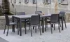 An outdoor dining set with a rectangular table and chairs placed on a paved patio in front of a stone building with a water wheel. Produktbild-Vorschau